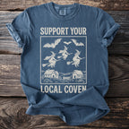 T-shirt with white graphic of witches flying on brooms over houses, and bats, reading "Support Your Local Coven".