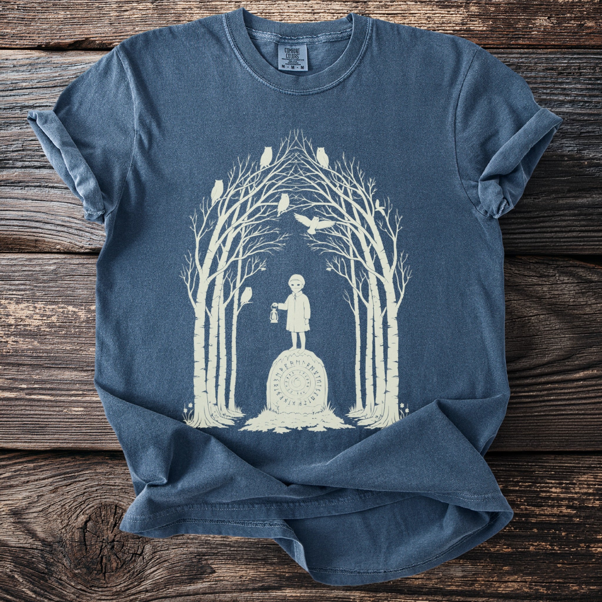 T-shirt with light beige graphic of a child holding a lantern on a stone, surrounded by trees and owls.