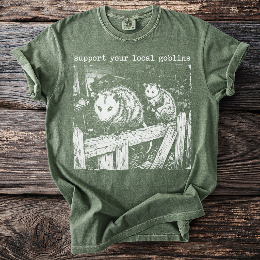 Green t-shirt with white graphic of two possums on a broken fence and text "support your local goblins".