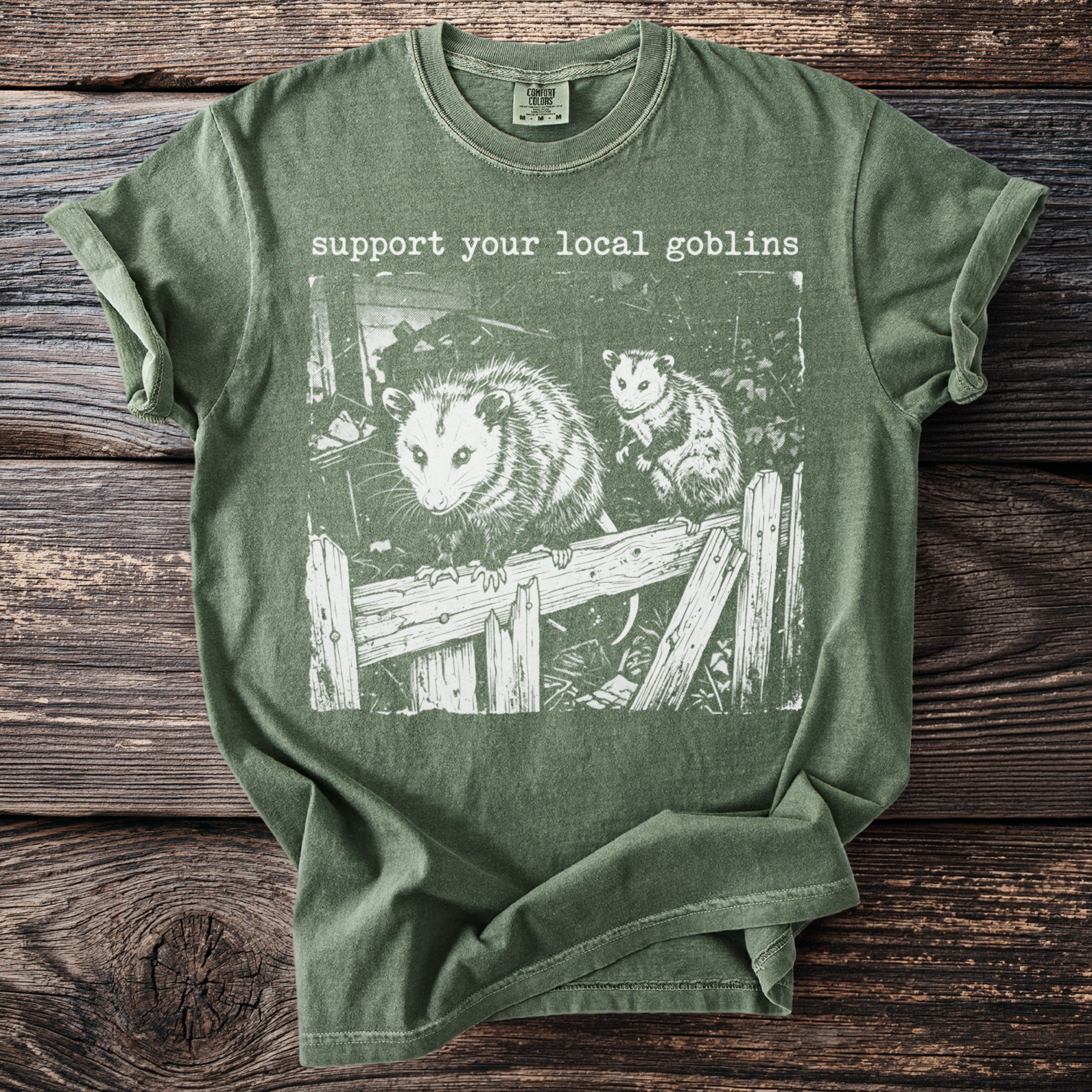 Green t-shirt with white graphic of two possums on a broken fence and text "support your local goblins".