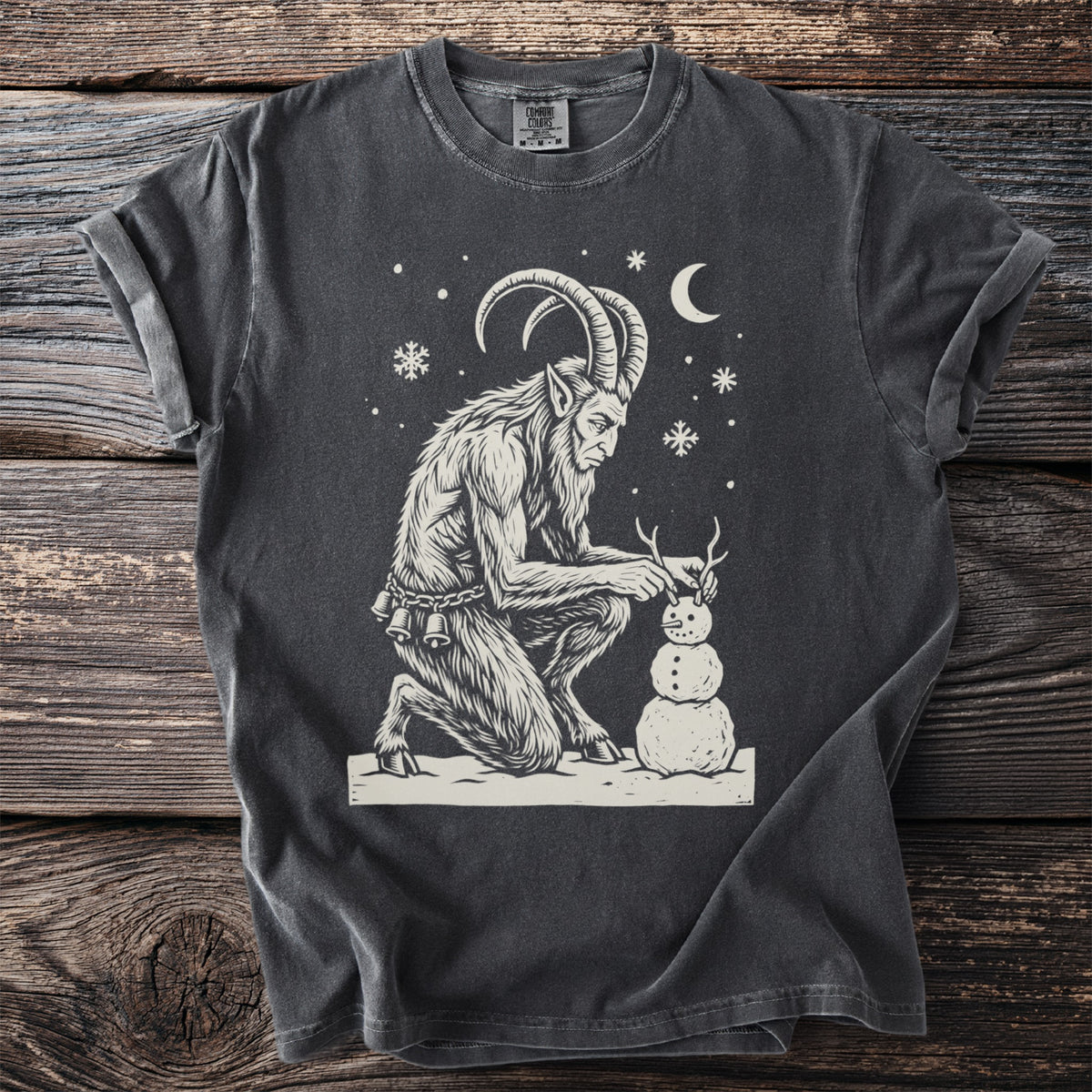 Pepper gray t-shirt with a cream folk-cut print: Krampus kneels in the snow, giving his snowman a pair of twig horns.