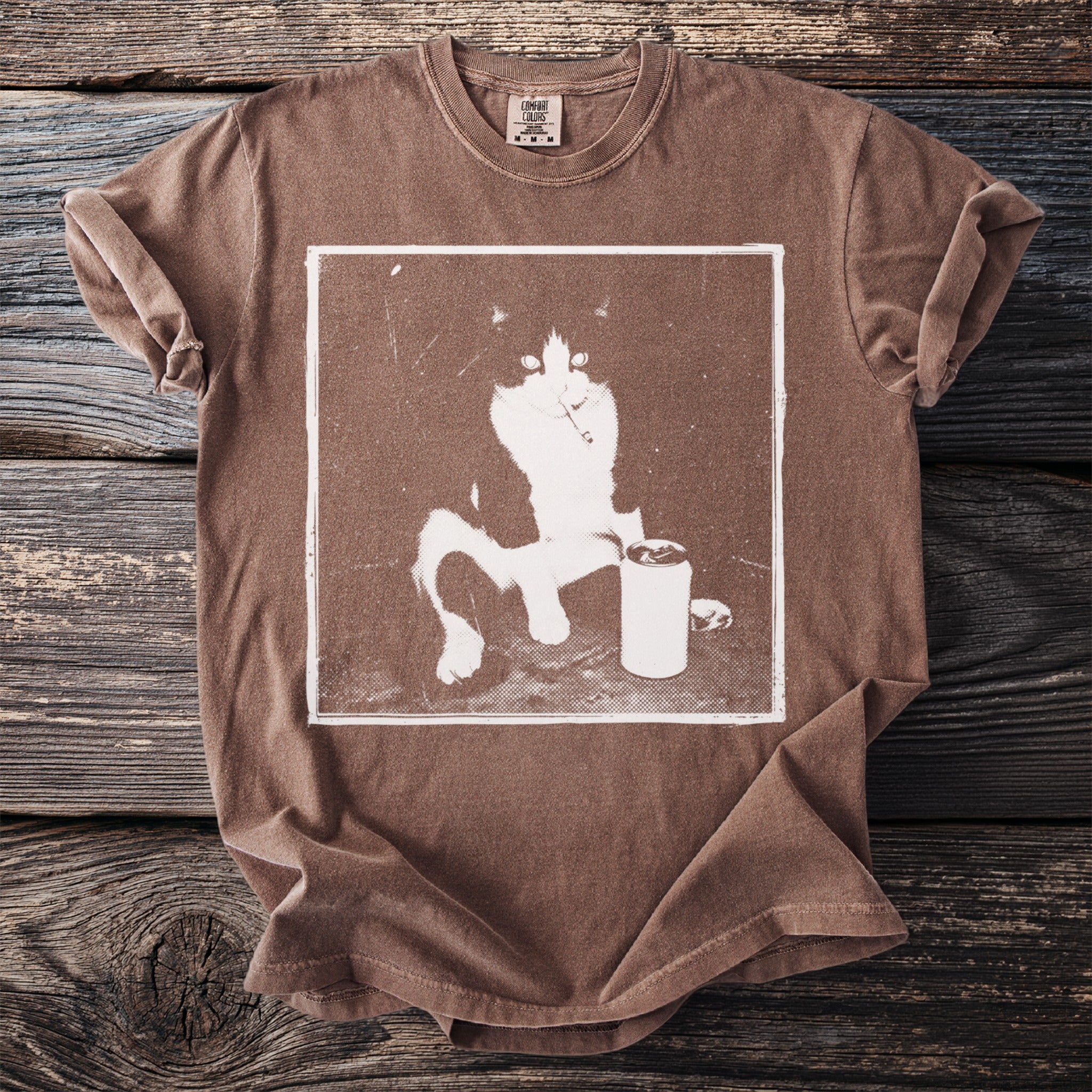 Brown t-shirt with a white graphic of a cat and a can on wood background.