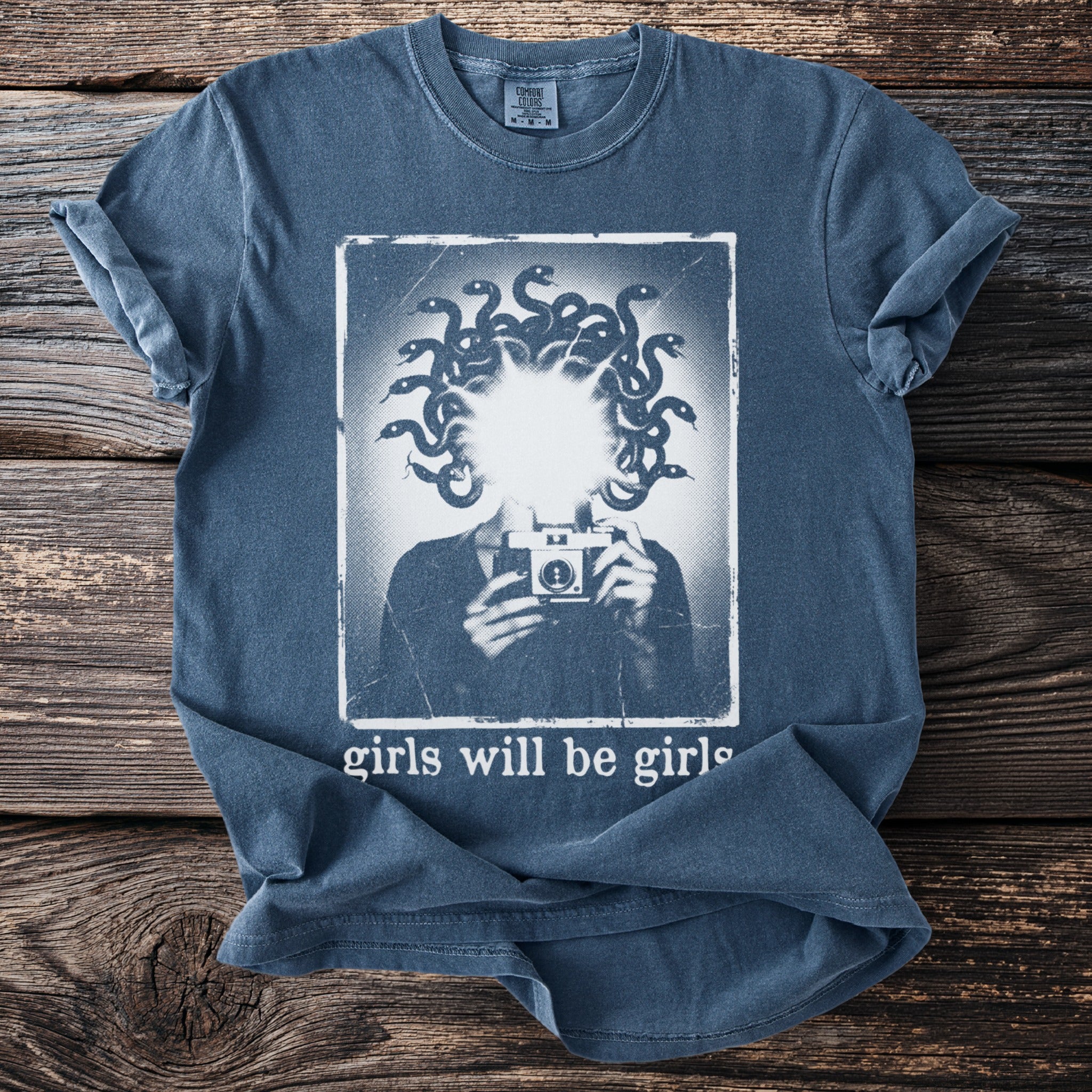 Blue t-shirt graphic of Medusa holding a camera with text "girls will be girls".