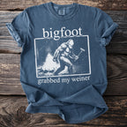Blue t-shirt with Bigfoot around a campfire holding a hot dog skewer.