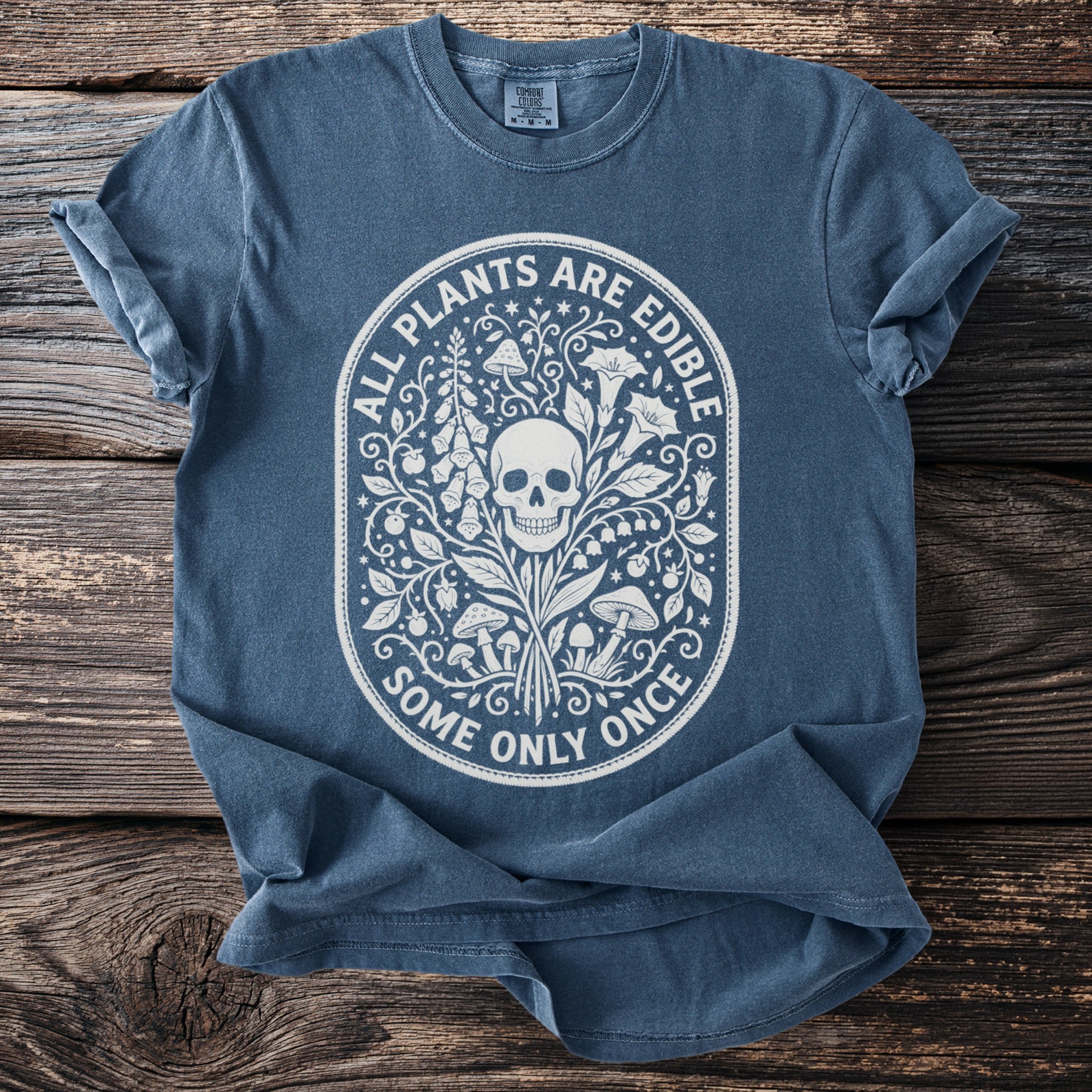 Blue t-shirt with white graphic of skull, plants, and mushrooms, text "ALL PLANTS ARE EDIBLE SOME ONLY ONCE".