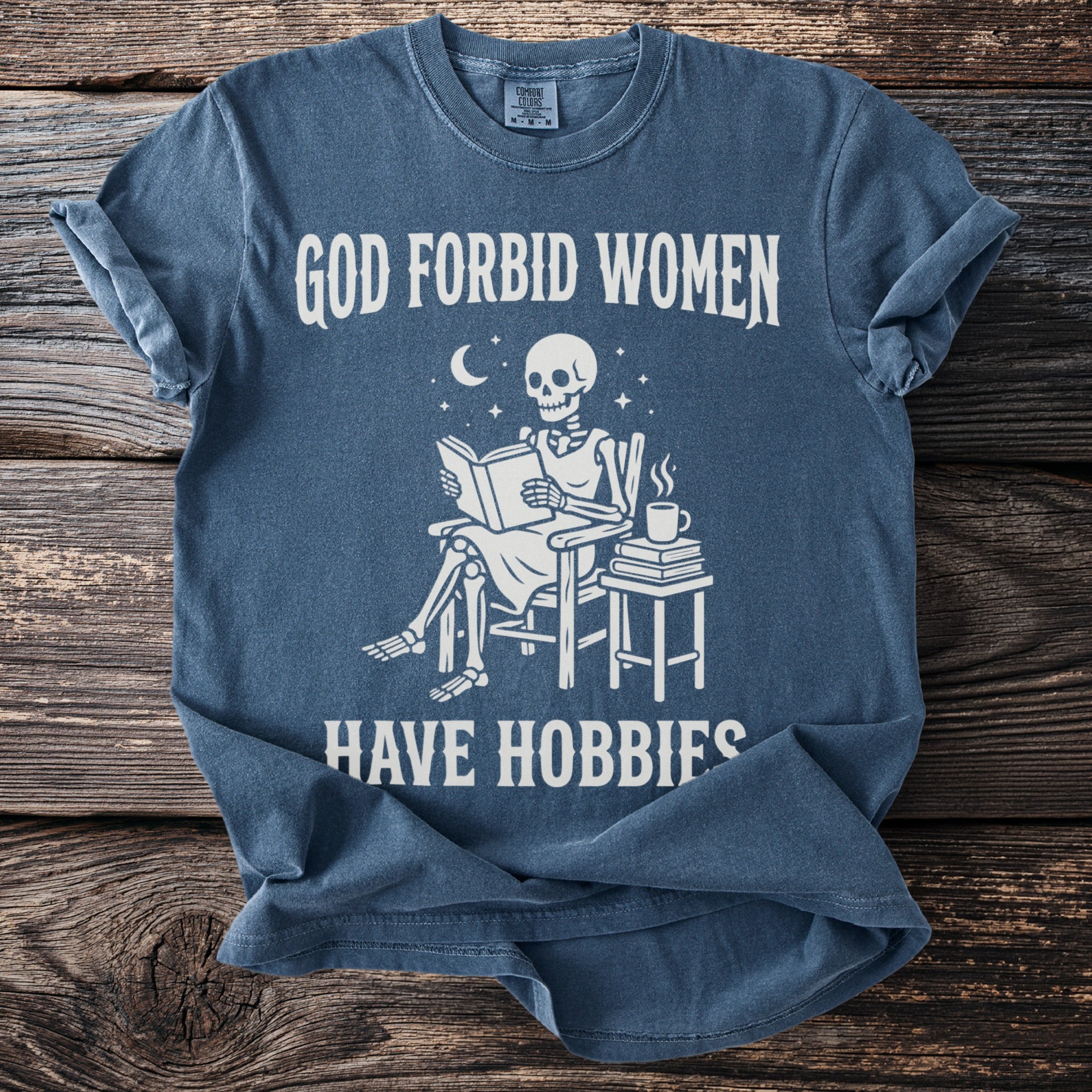 Blue t-shirt with skeleton reading a book and text: God forbid women have hobbies.