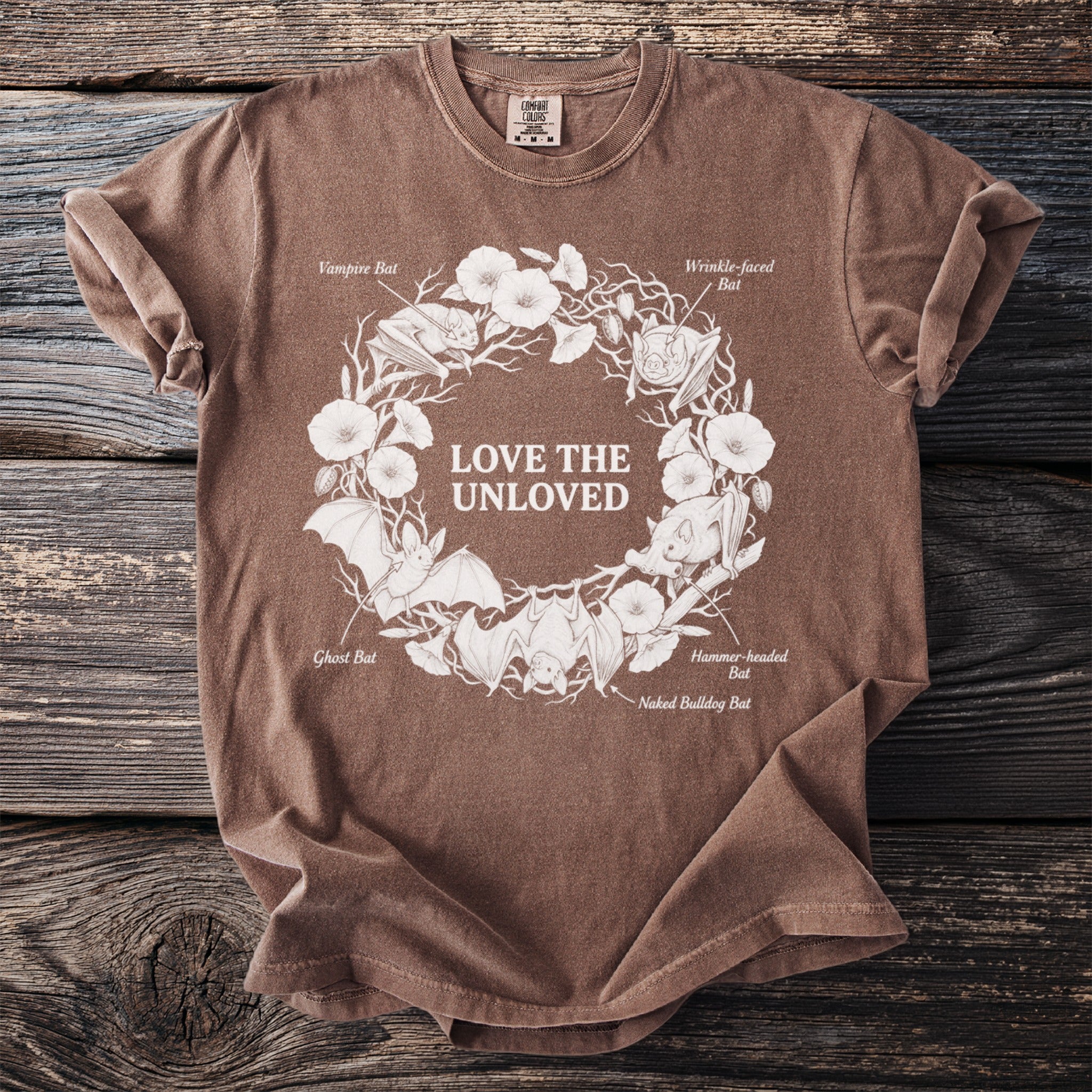 Brown t-shirt with white illustration of bats, flowers, and text "LOVE THE UNLOVED".