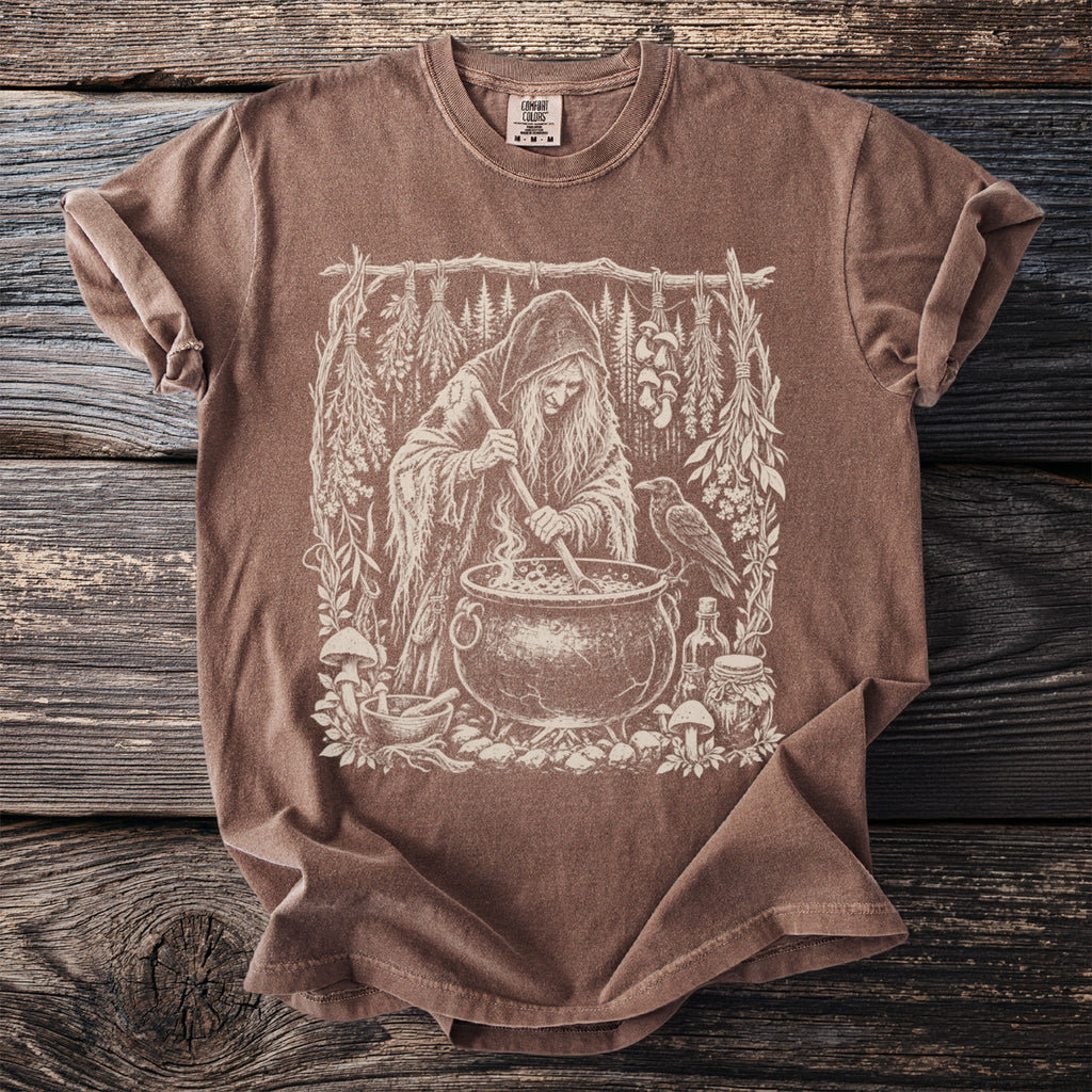 Brown t-shirt with a vintage sketch of a witch stirring a cauldron with a raven nearby.
