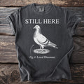 Dark gray t-shirt with "STILL HERE" above a pigeon labeled "Fig. 1: Local Dinosaur."