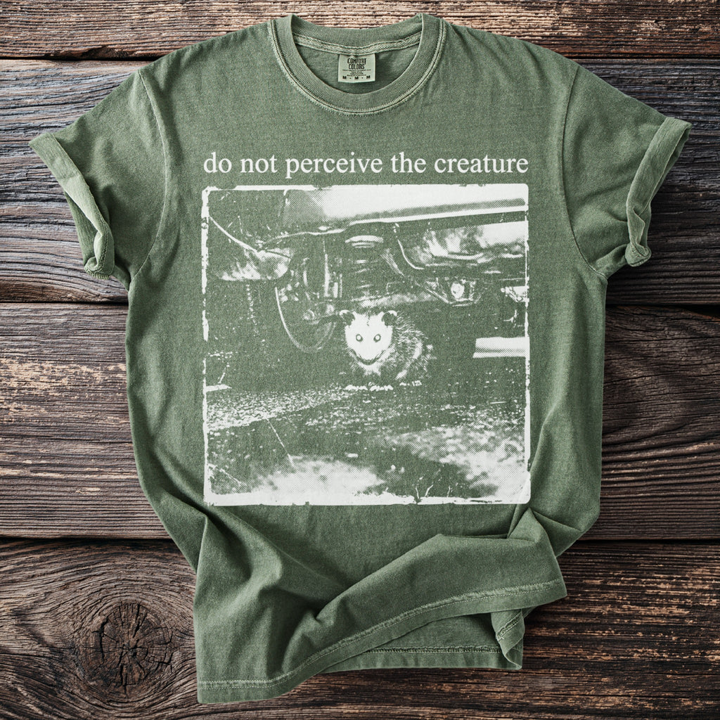 Green t-shirt with white graphic of creature under car and text "do not perceive the creature".
