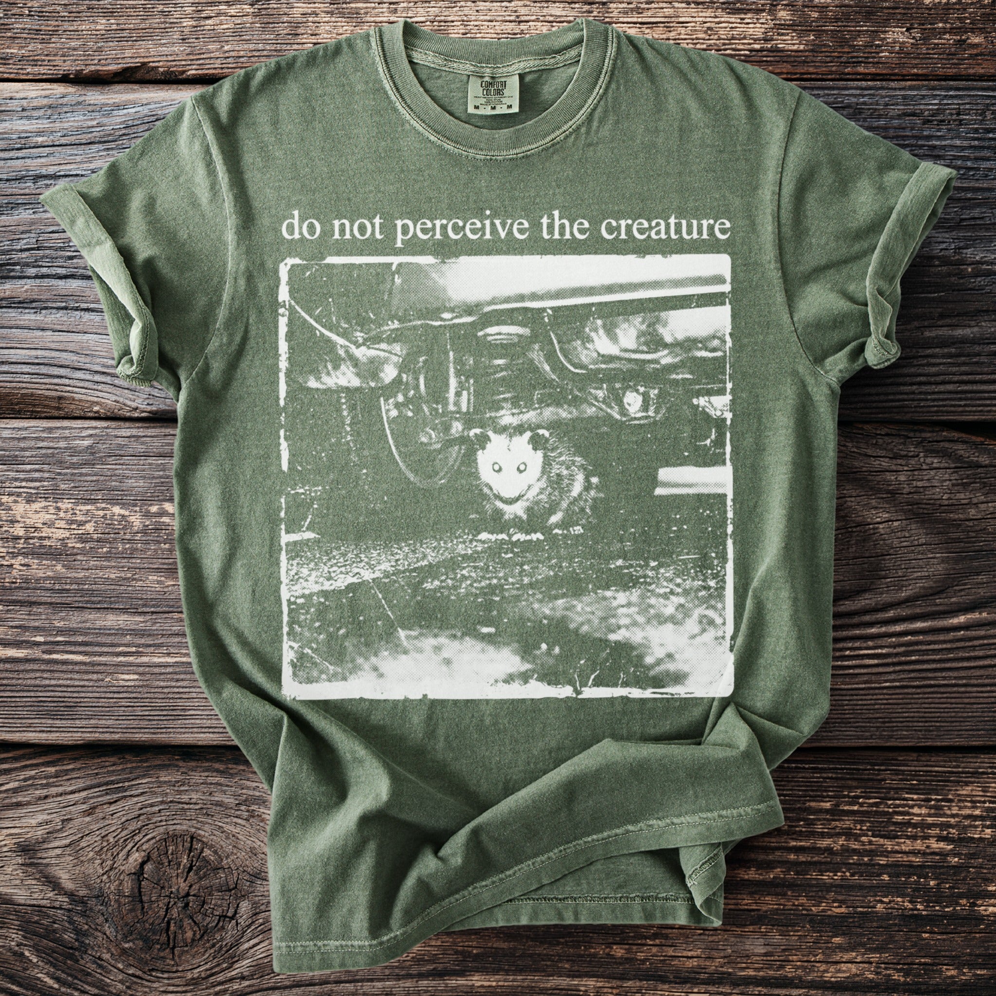 Green t-shirt with white graphic of creature under car and text "do not perceive the creature".