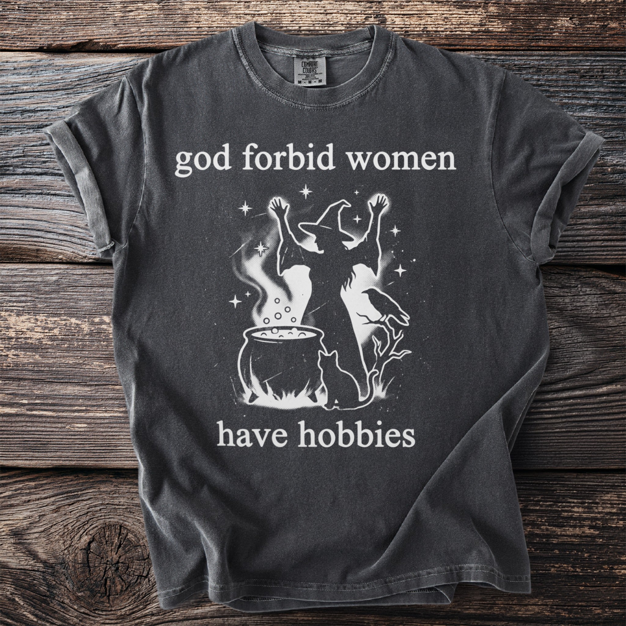 Dark gray t-shirt with white text "god forbid women have hobbies" and witch graphic.