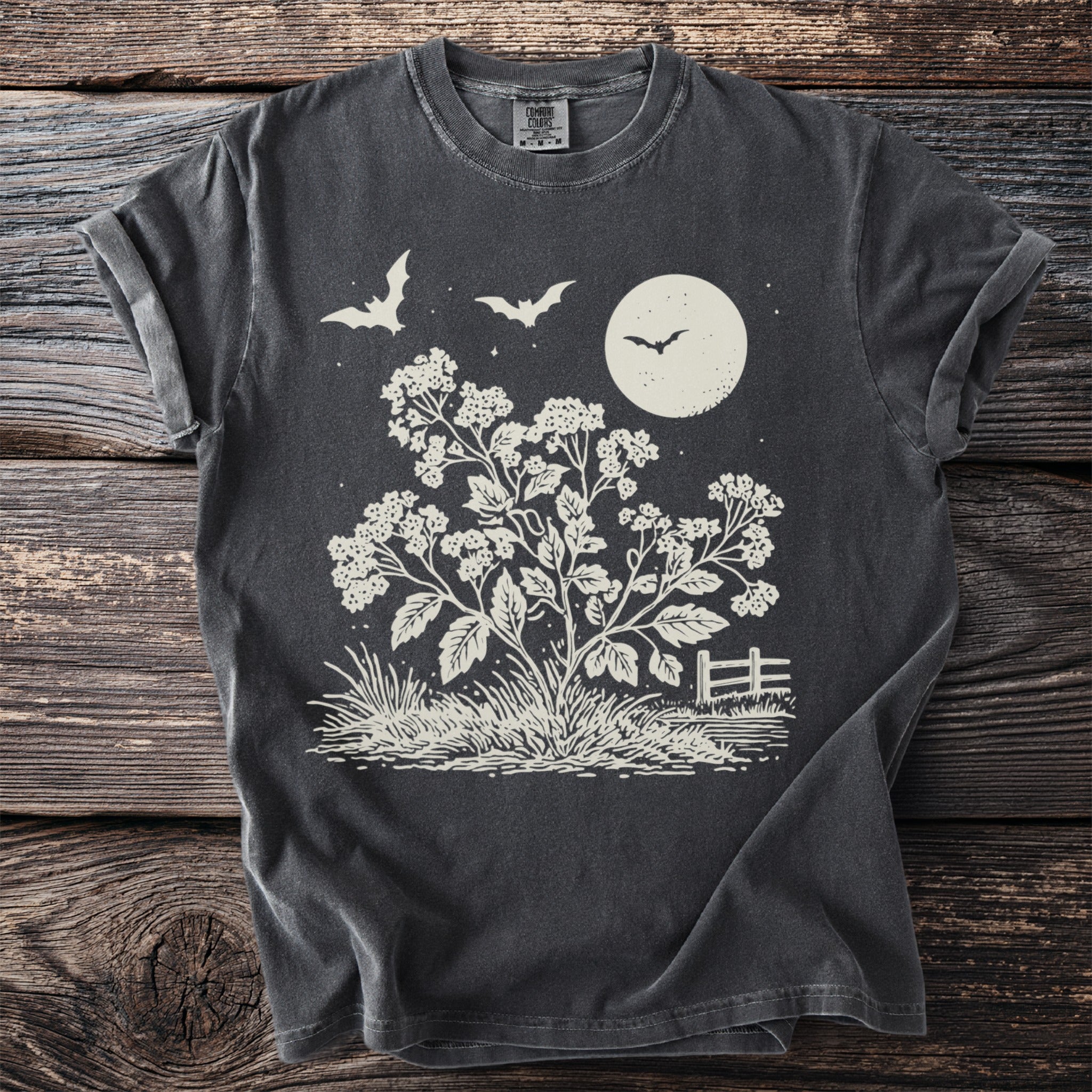 Dark t-shirt with a cream graphic of bats, moon, flowering plant, and fence.