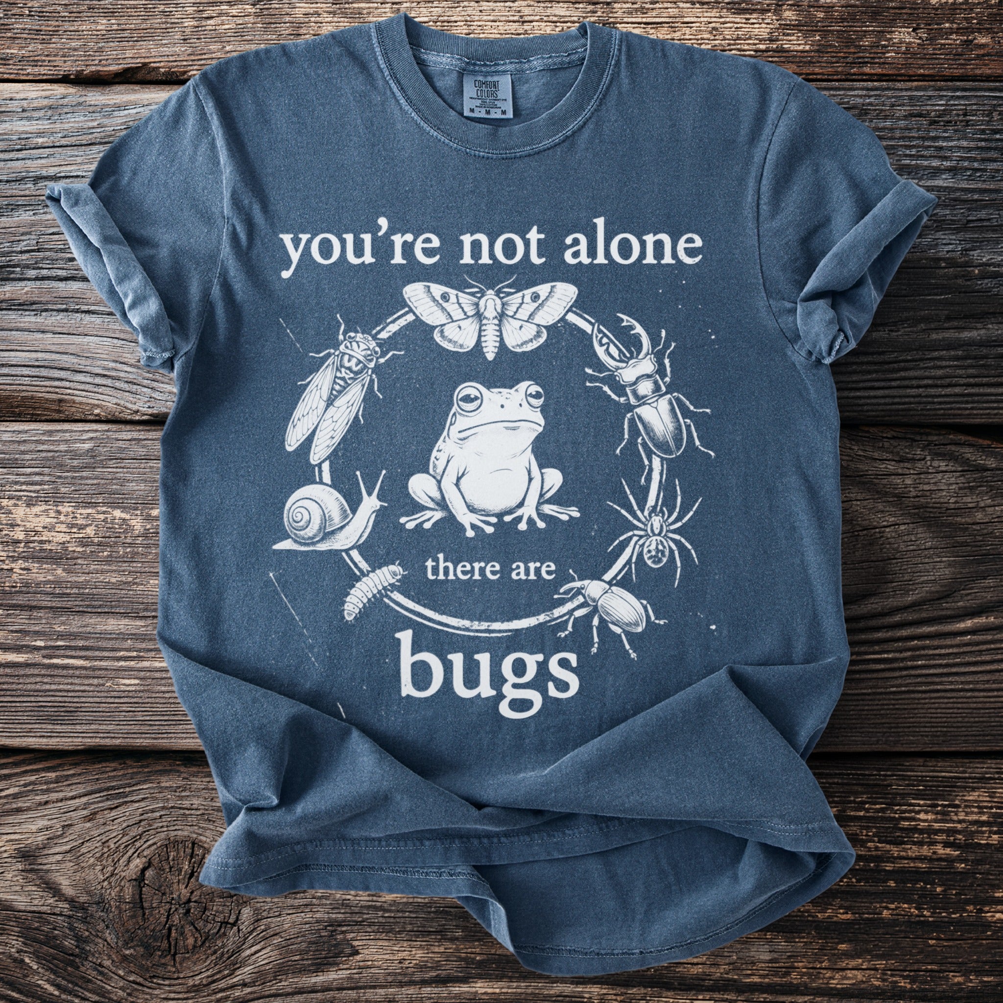 Blue t-shirt with a circular design of bugs and insects surrounding a frog, reading "you're not alone there are bugs".