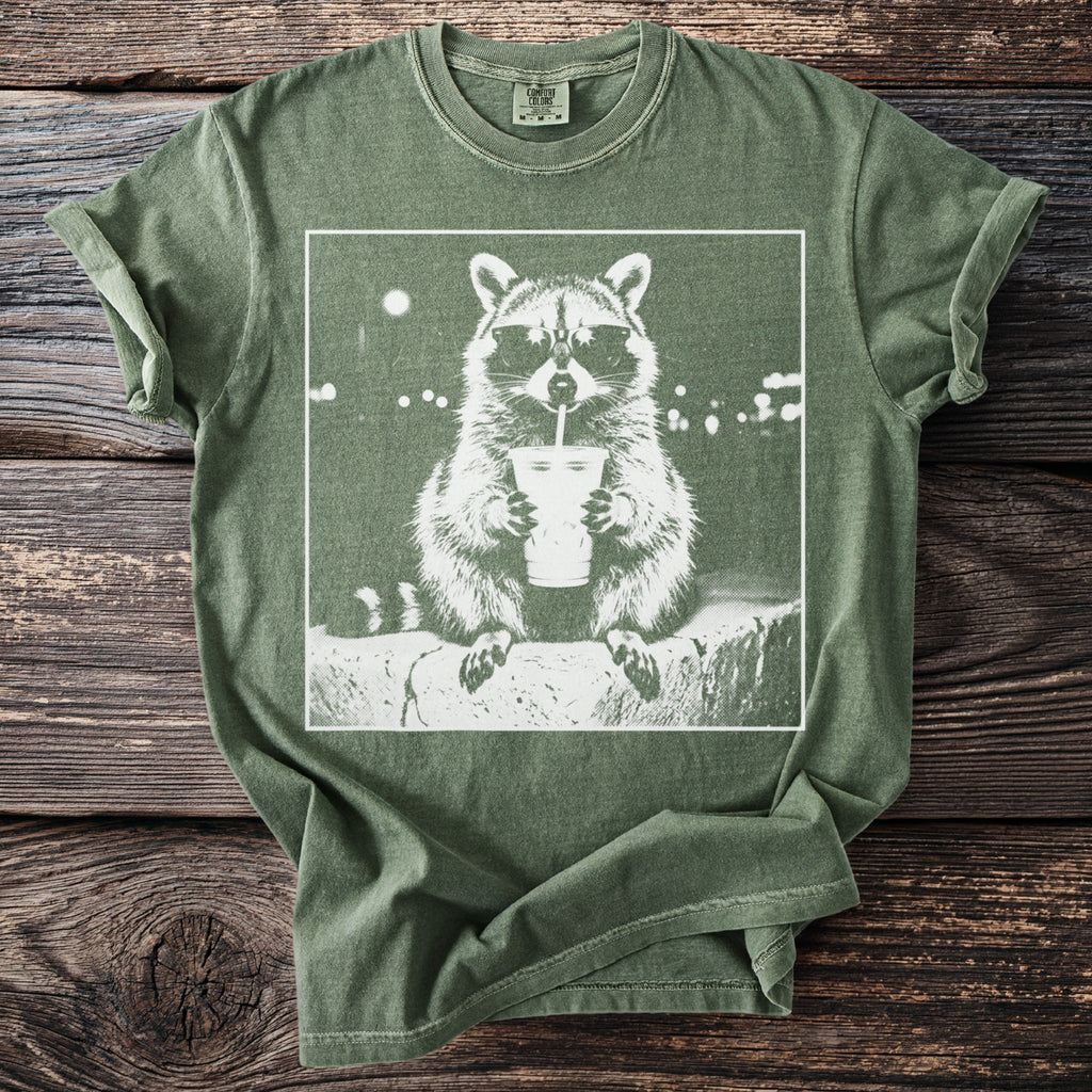 Green t-shirt with white graphic of a raccoon wearing sunglasses and drinking from a cup.