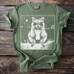 Green t-shirt with white graphic of a raccoon wearing sunglasses and drinking from a cup.