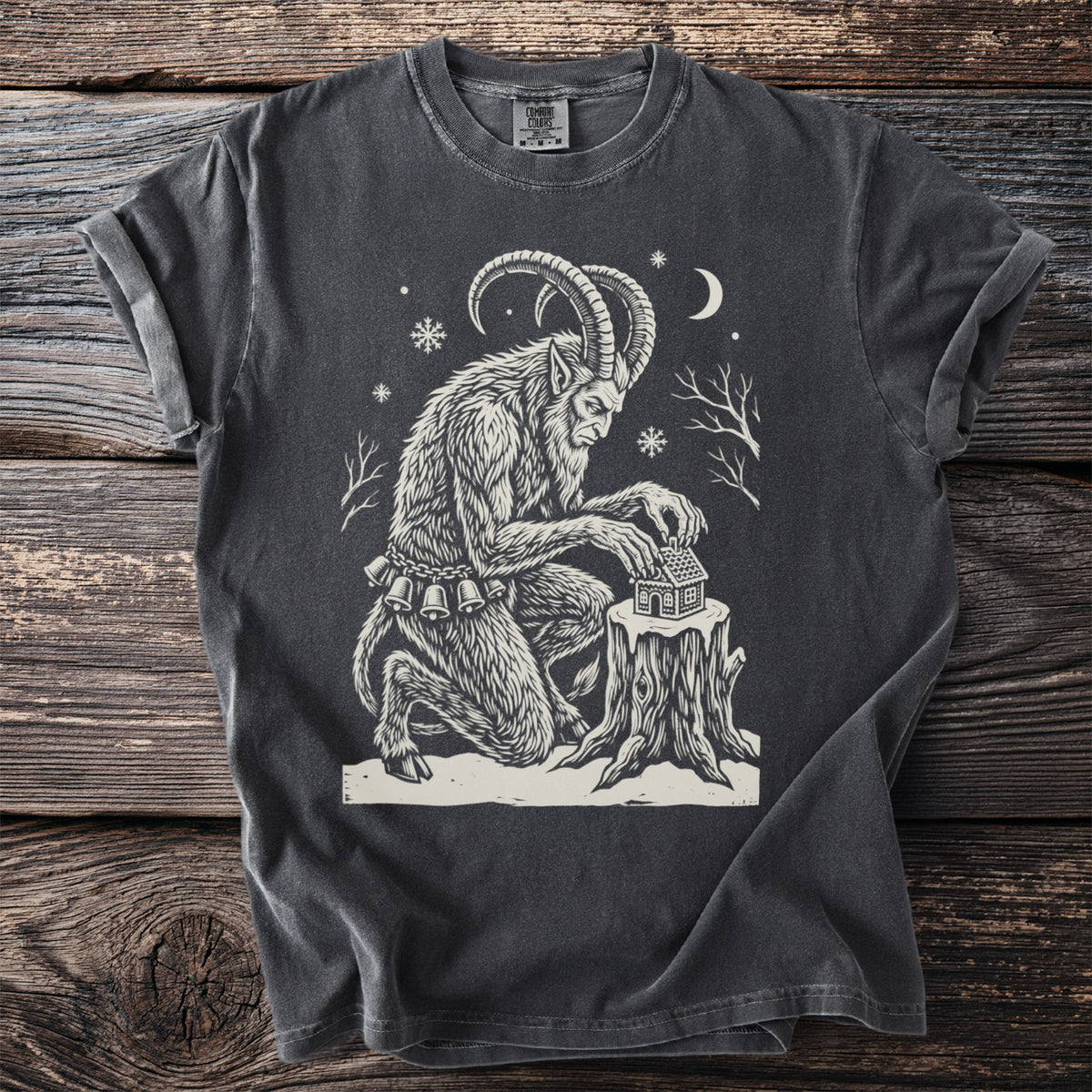 Pepper gray t-shirt with a cream folk-cut print: Krampus kneels by a tree stump in the snow, setting a tiny gingerbread house on top.