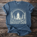 Blue t-shirt with print of Bigfoot, a small figure, lake, and forest under starry sky.