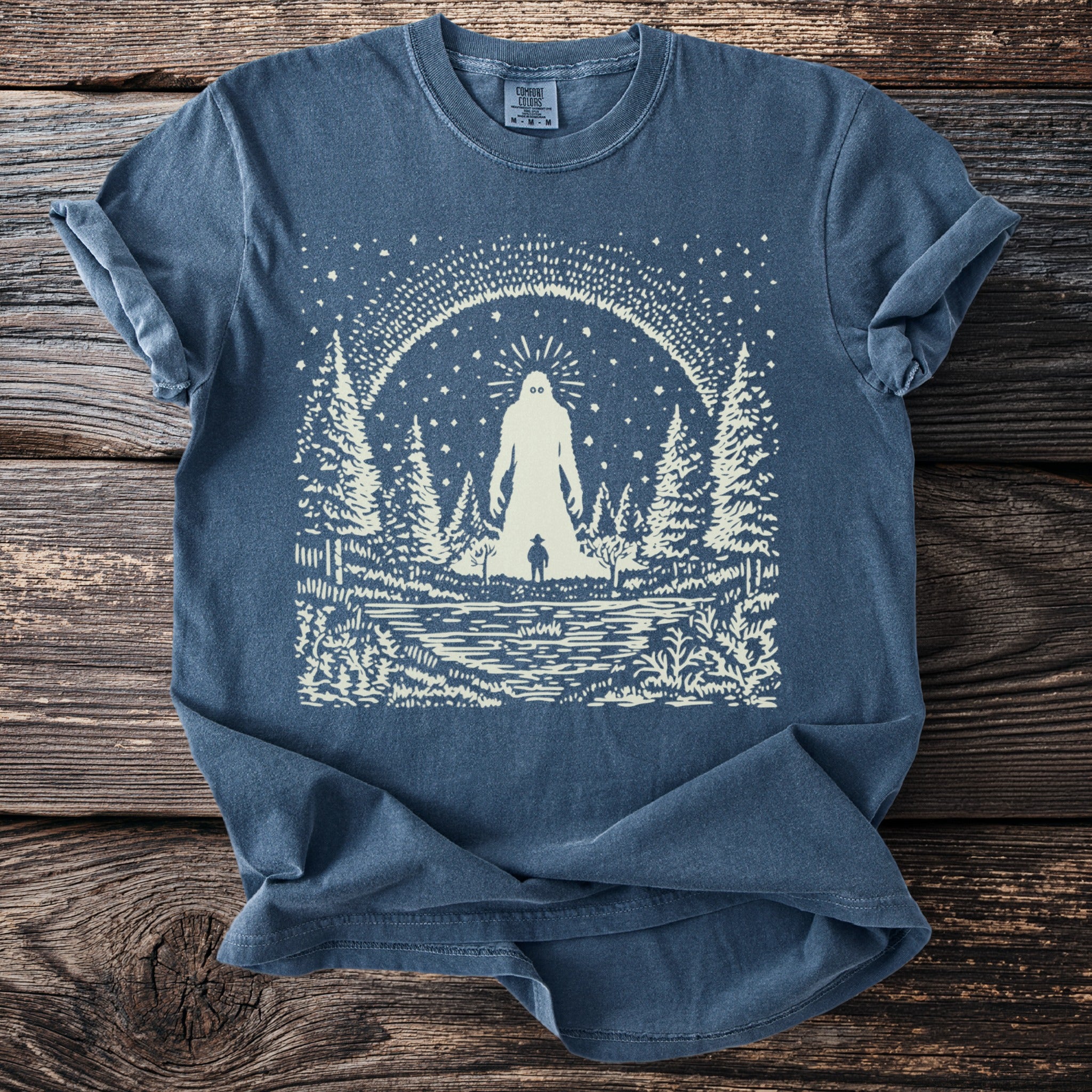 Blue t-shirt with print of Bigfoot, a small figure, lake, and forest under starry sky.