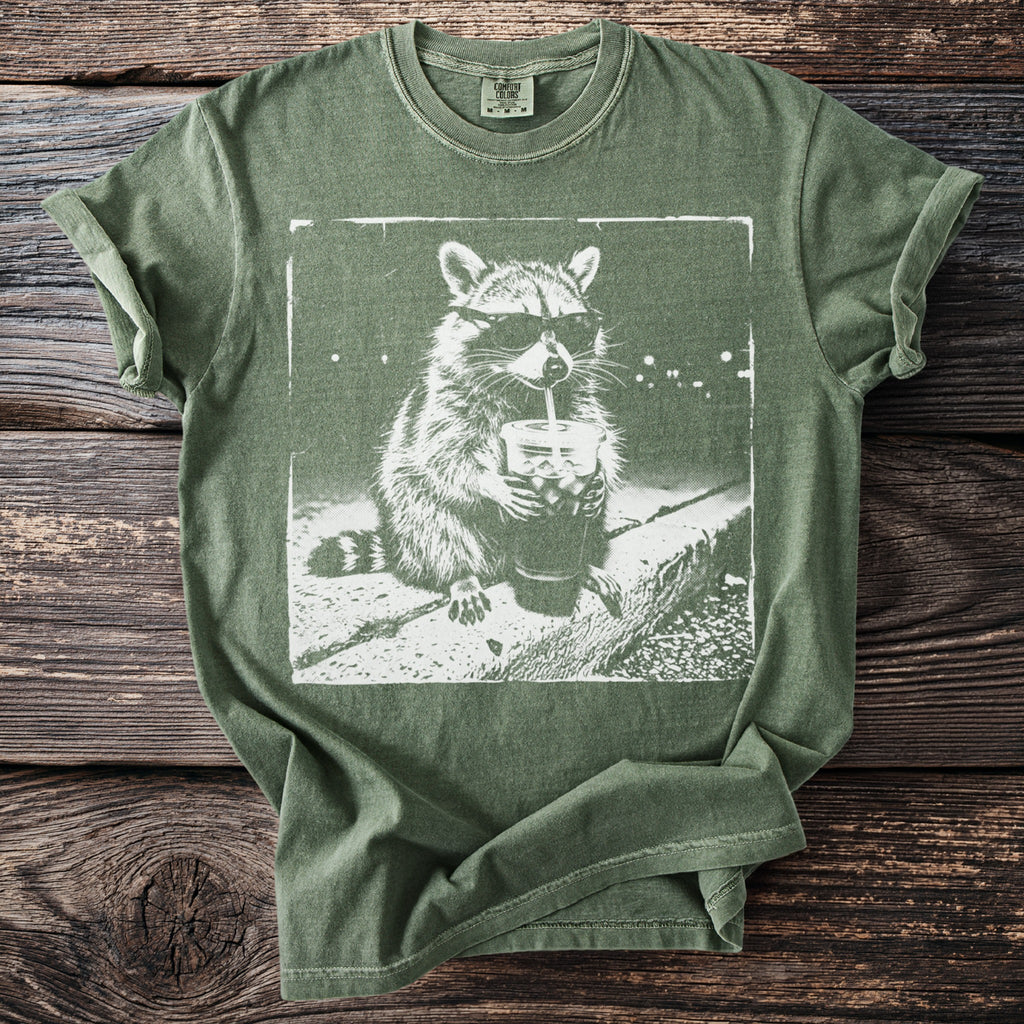 Green t-shirt with white graphic of a raccoon wearing sunglasses drinking from a cup.