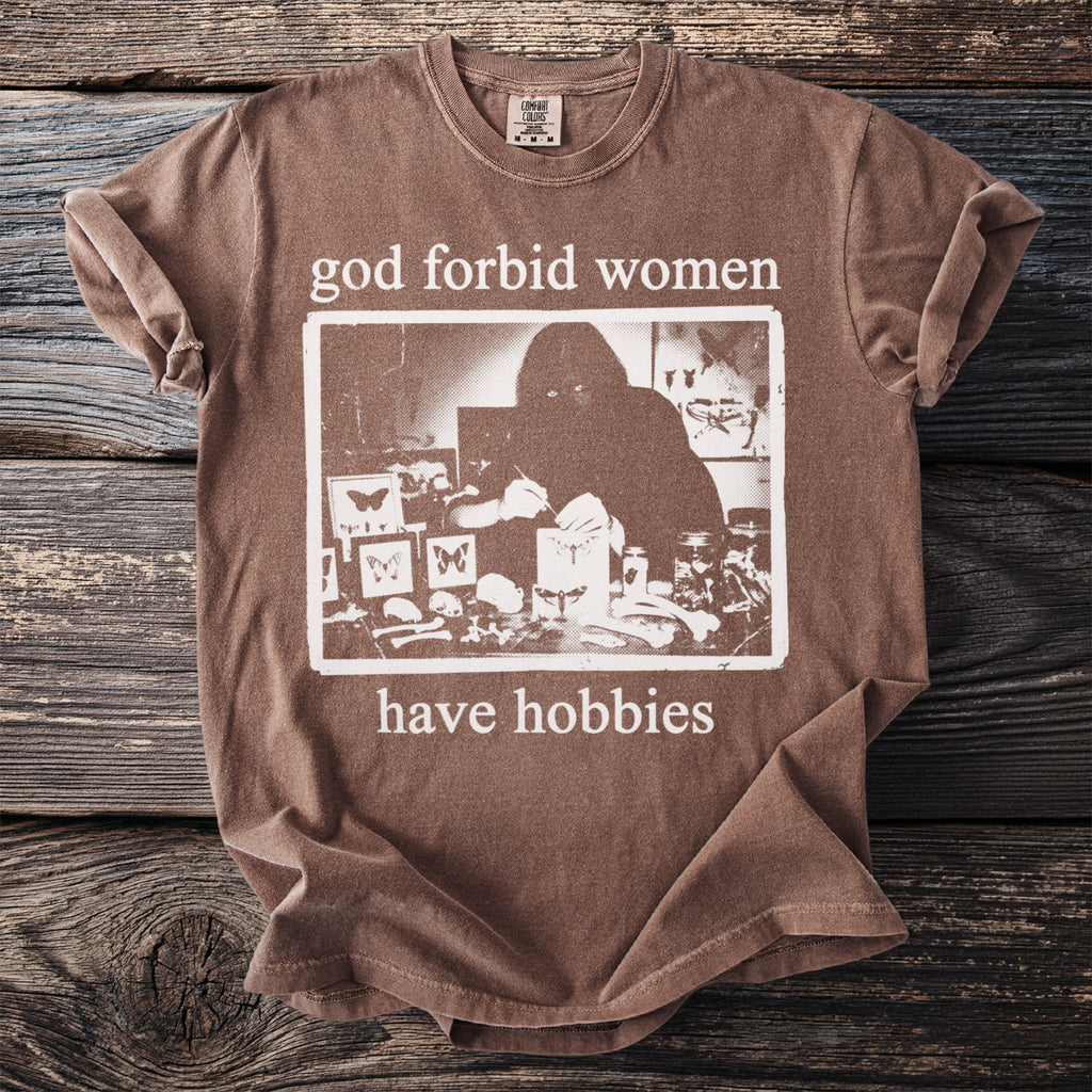 Brown t-shirt with "god forbid women have hobbies" text and a graphic of a person examining butterflies.