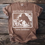 Brown t-shirt with "god forbid women have hobbies" text and a graphic of a person examining butterflies.