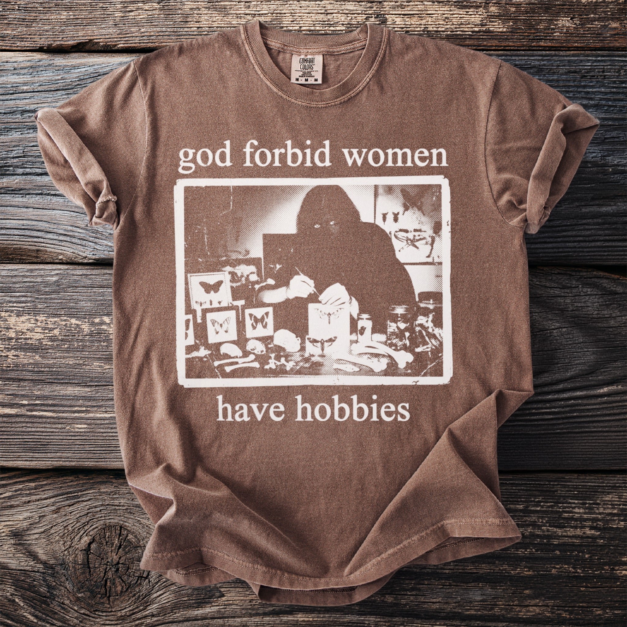 Brown t-shirt with "god forbid women have hobbies" text and a graphic of a person examining butterflies.