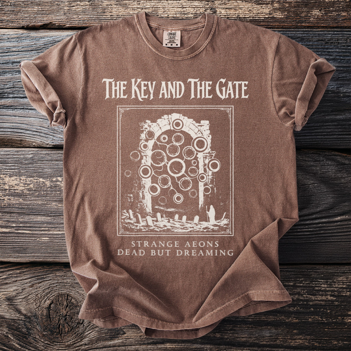 Brown t-shirt with "The Key and the Gate" graphic, "Strange Aeons Dead But Dreaming" text.