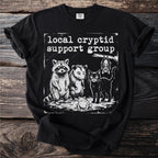 Black t-shirt with "Local Cryptid Support Group" text and raccoon, opossum, cat, frog, and bat illustration.