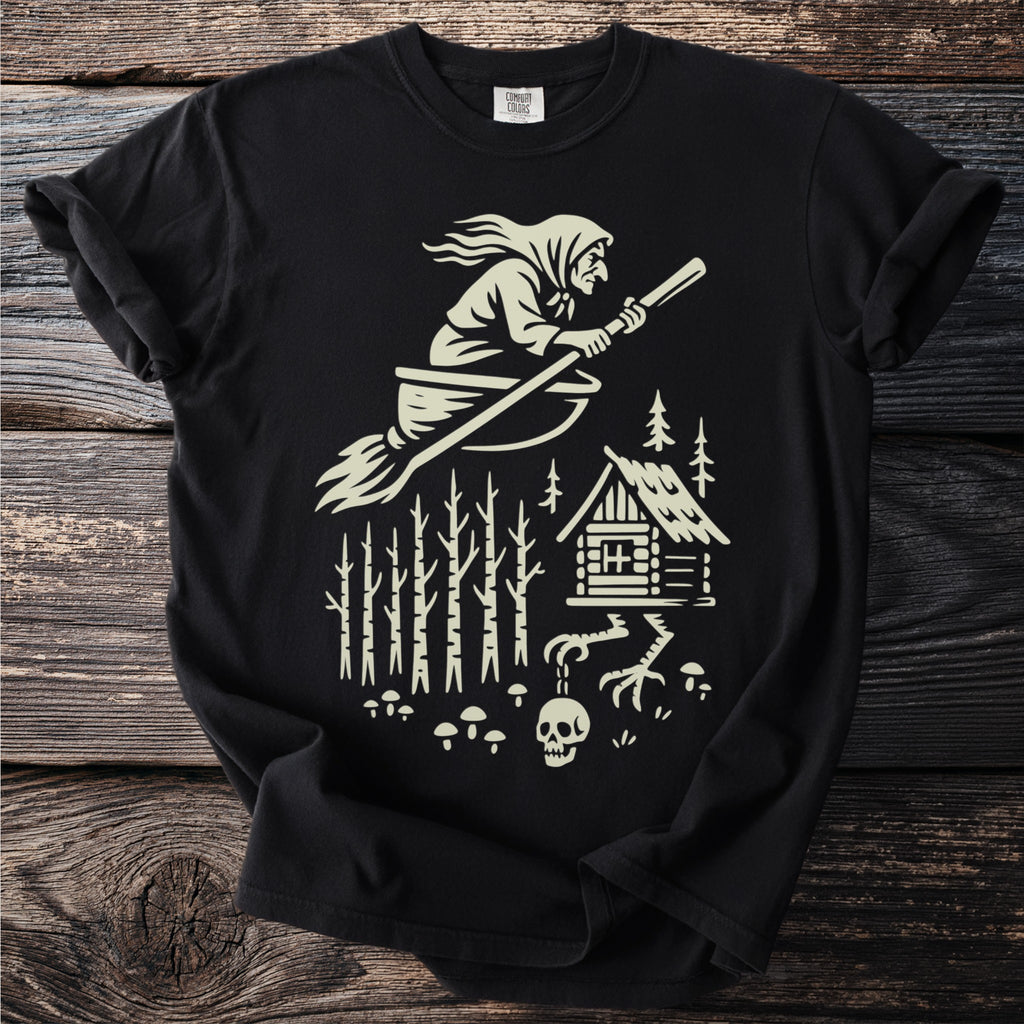 Black t-shirt with a graphic of a witch on a broomstick flying over a forest and cabin.