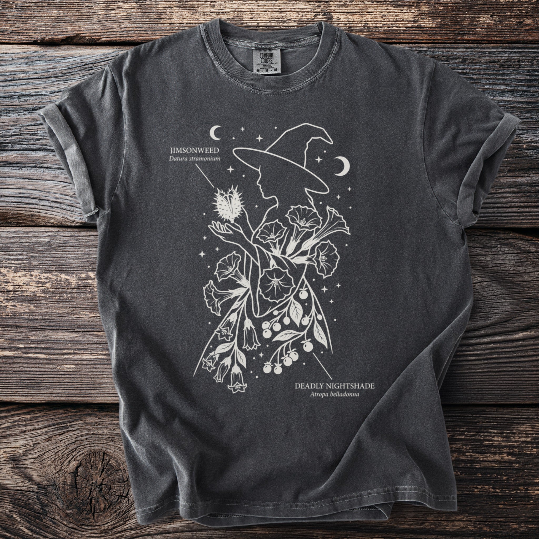 Dark t-shirt with white line art of a witch holding Jimsonweed and Deadly Nightshade flowers.