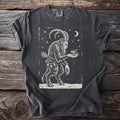 Pepper gray t-shirt with a cream folk-cut print: Krampus stands in the snow, birch switches at his back, holding a small songbird in his hand.