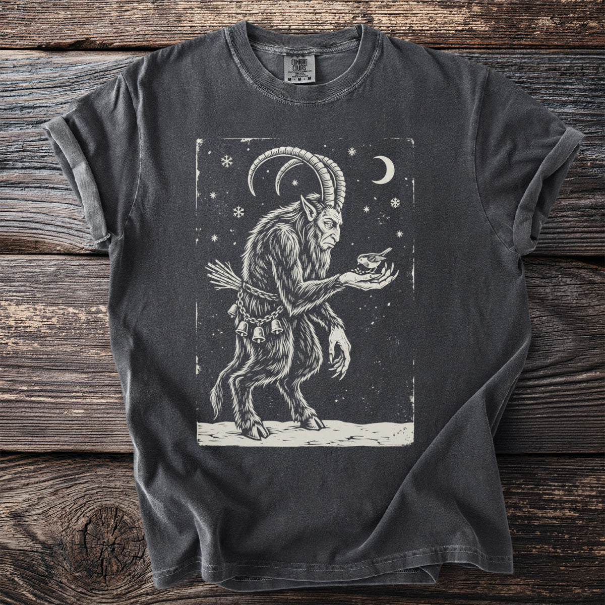 Pepper gray t-shirt with a cream folk-cut print: Krampus stands in the snow, birch switches at his back, holding a small songbird in his hand.