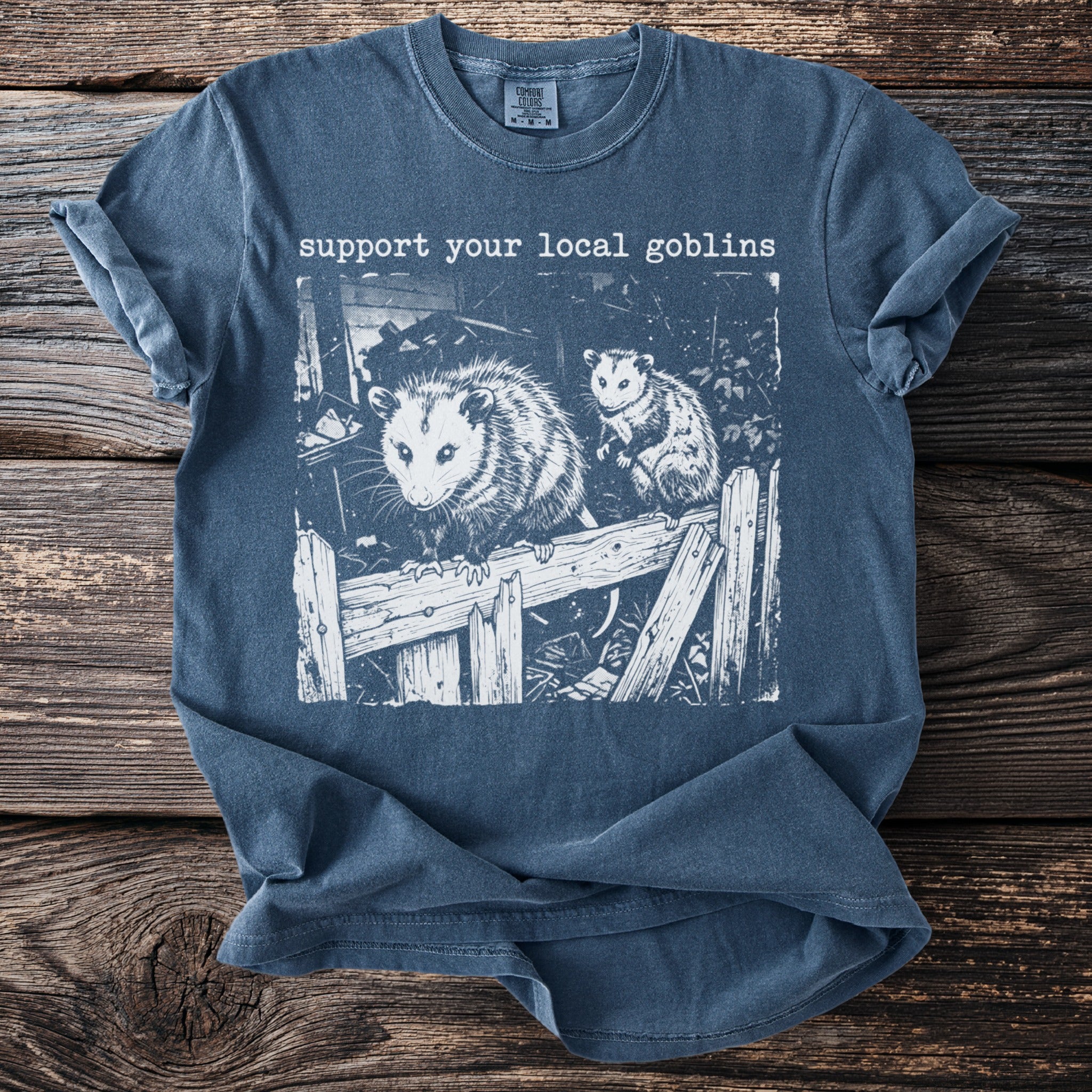 Blue t-shirt with two opossums on a wooden fence and text "support your local goblins".