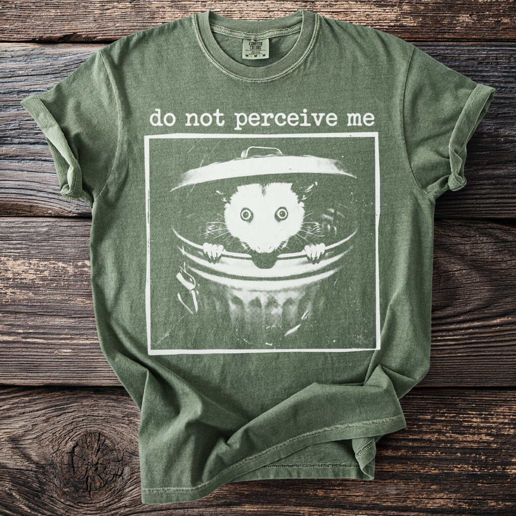 Olive green t-shirt with "do not perceive me" and opossum in trash can graphic.