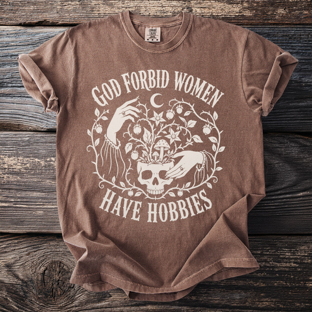 Brown t-shirt with white text "GOD FORBID WOMEN HAVE HOBBIES" alongside a skull and floral design.