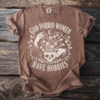 Brown t-shirt with white text "GOD FORBID WOMEN HAVE HOBBIES" alongside a skull and floral design.
