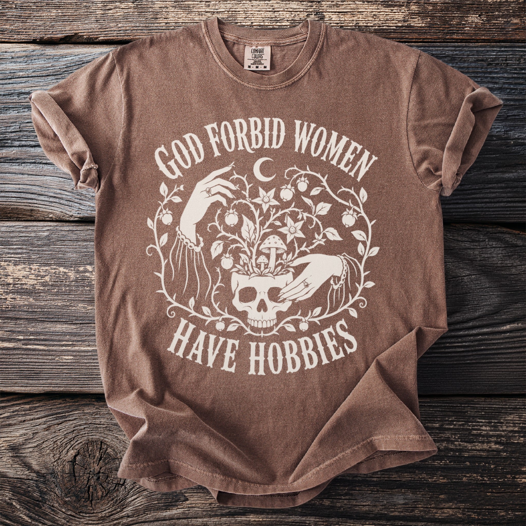 Brown t-shirt with white text "GOD FORBID WOMEN HAVE HOBBIES" alongside a skull and floral design.
