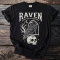 Black t-shirt with white graphic of a raven on a skull, text "Raven gothic crow corvid familiar".