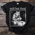 Black t-shirt with "soft but feral" text and a werewolf with a kitten in a box.