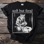 Black t-shirt with "soft but feral" text and a werewolf with a kitten in a box.