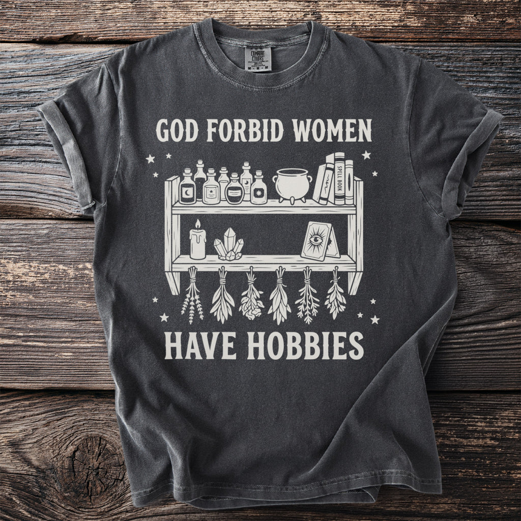 Dark gray t-shirt with white graphic of potions, crystals, and herbs, reading "God Forbid Women Have Hobbies."