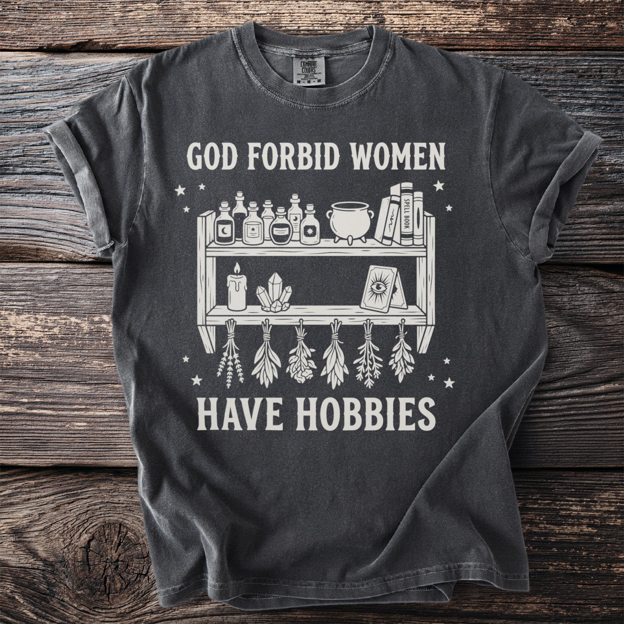 Dark gray t-shirt with white graphic of potions, crystals, and herbs, reading "God Forbid Women Have Hobbies."