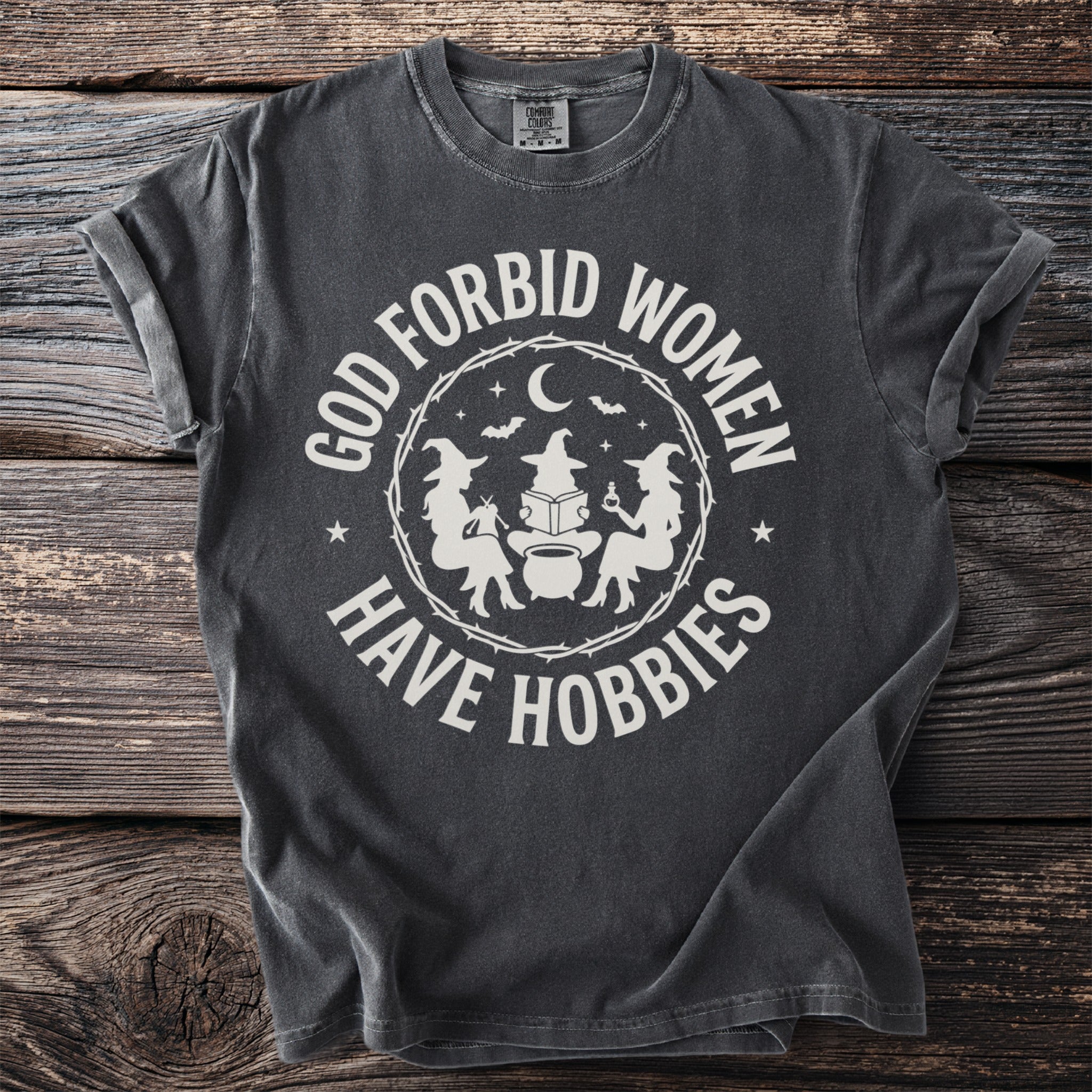 Dark gray t-shirt with white text "GOD FORBID WOMEN HAVE HOBBIES" and witch graphics.