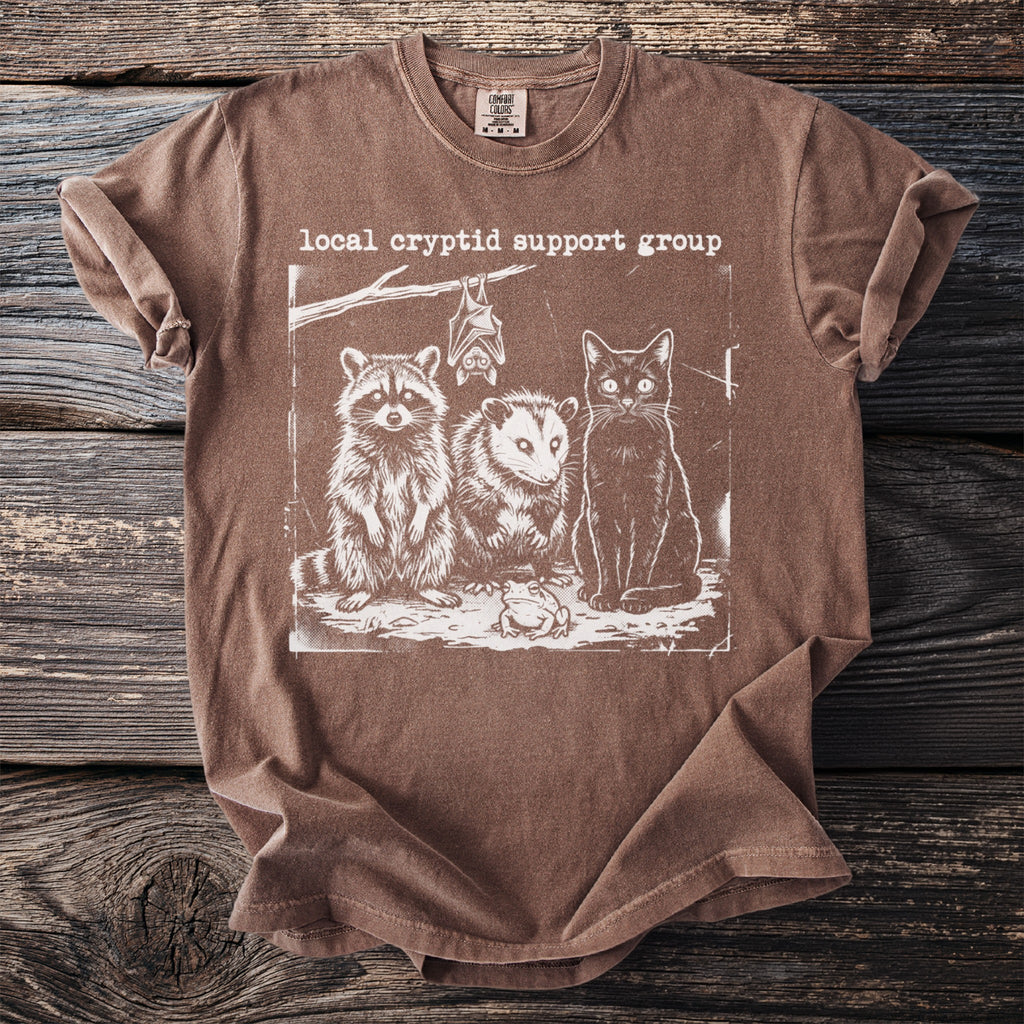 Brown t-shirt with "local cryptid support group" text and raccoon, opossum, cat, bat, and frog illustration.