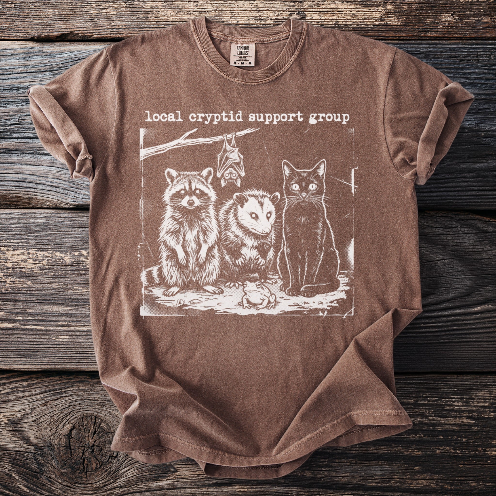 Brown t-shirt with "local cryptid support group" text and raccoon, opossum, cat, bat, and frog illustration.