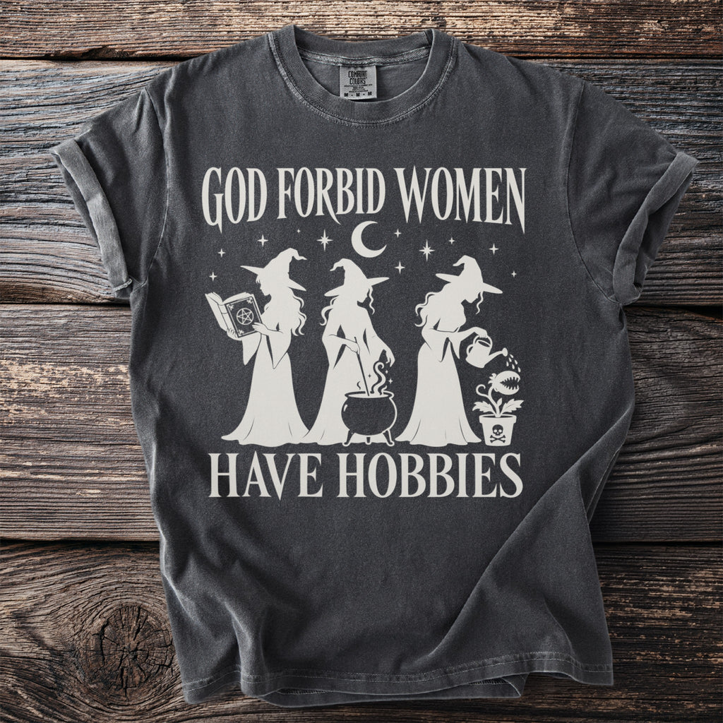 Dark t-shirt with cream graphic of three witches and text "GOD FORBID WOMEN HAVE HOBBIES".