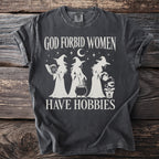 Dark t-shirt with cream graphic of three witches and text "GOD FORBID WOMEN HAVE HOBBIES".