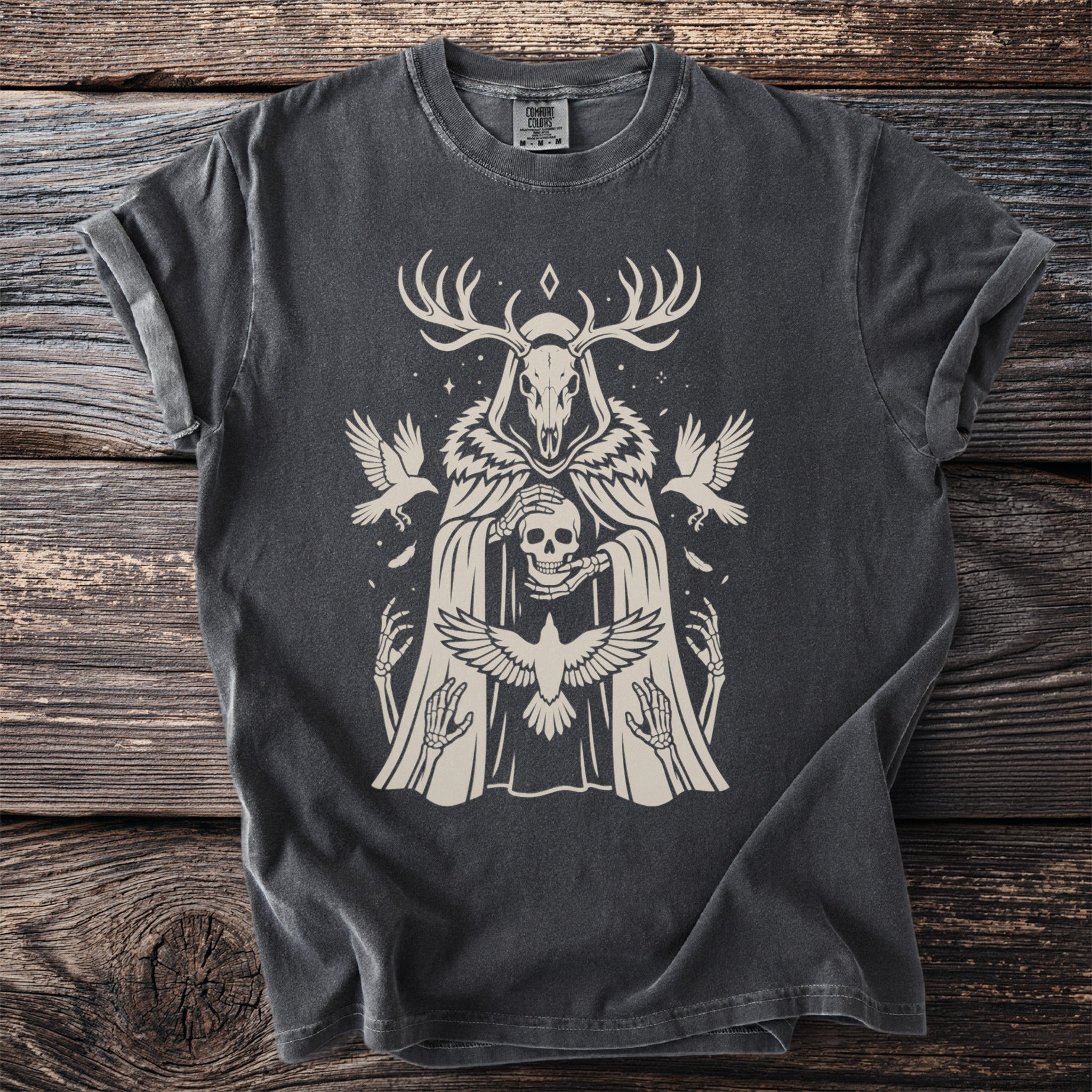 Dark t-shirt with cream graphic of a figure in a hooded robe holding a skull, with birds and antlers.