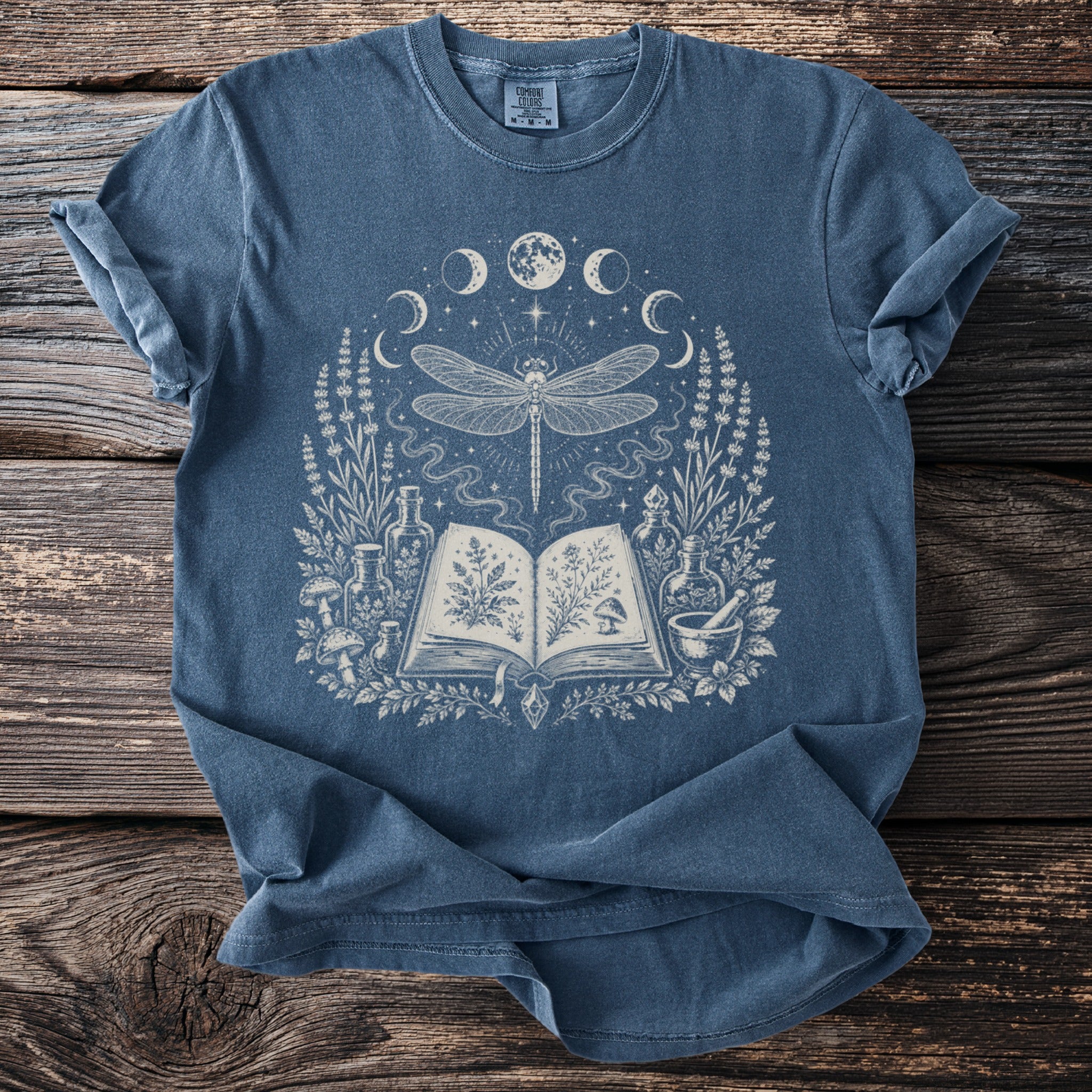 Blue t-shirt with a white graphic of a dragonfly, moon phases, and botanical elements.